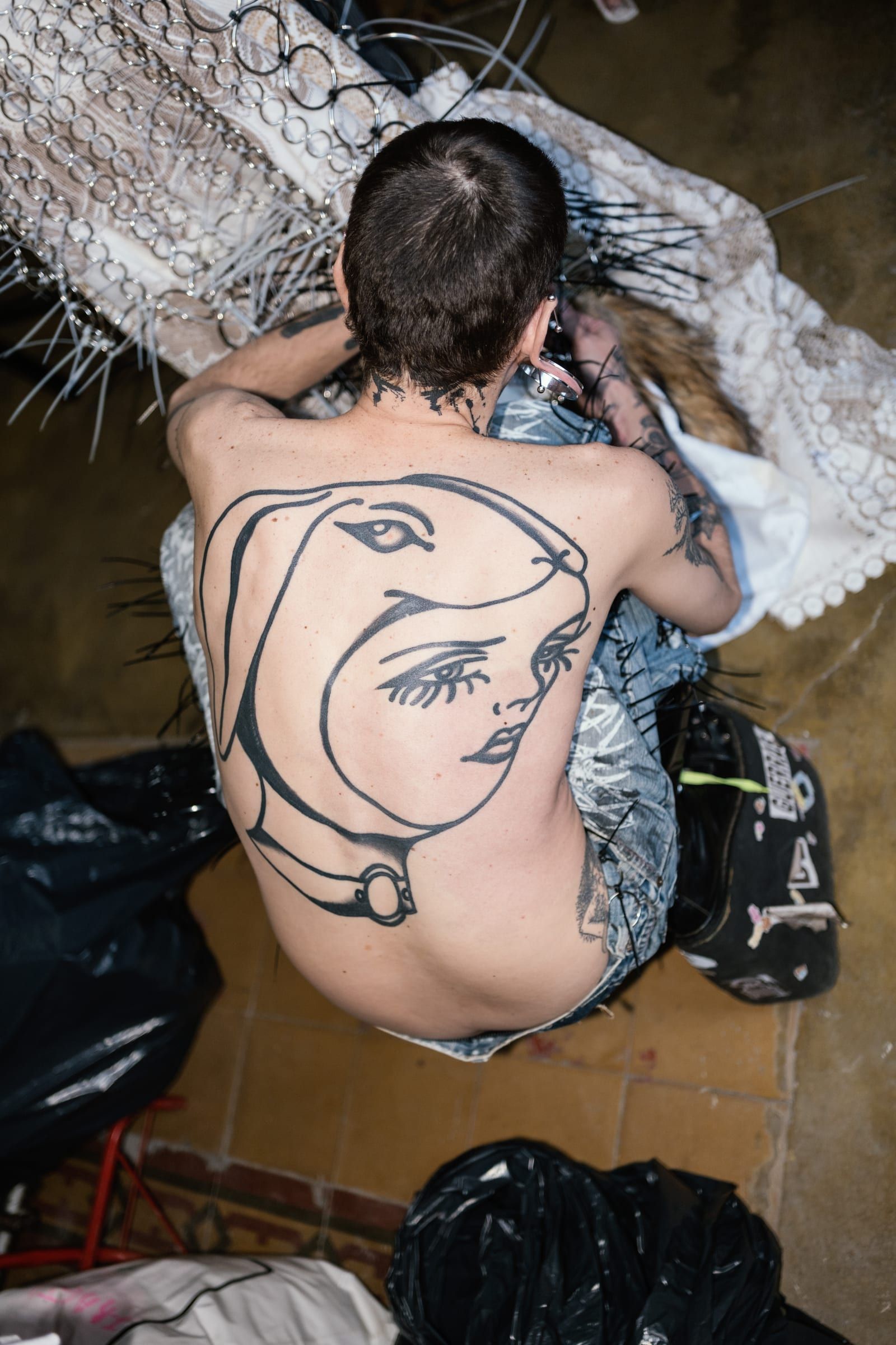 A bare back with a face drawn in ink, as the person gets dressed sitting on the floor.