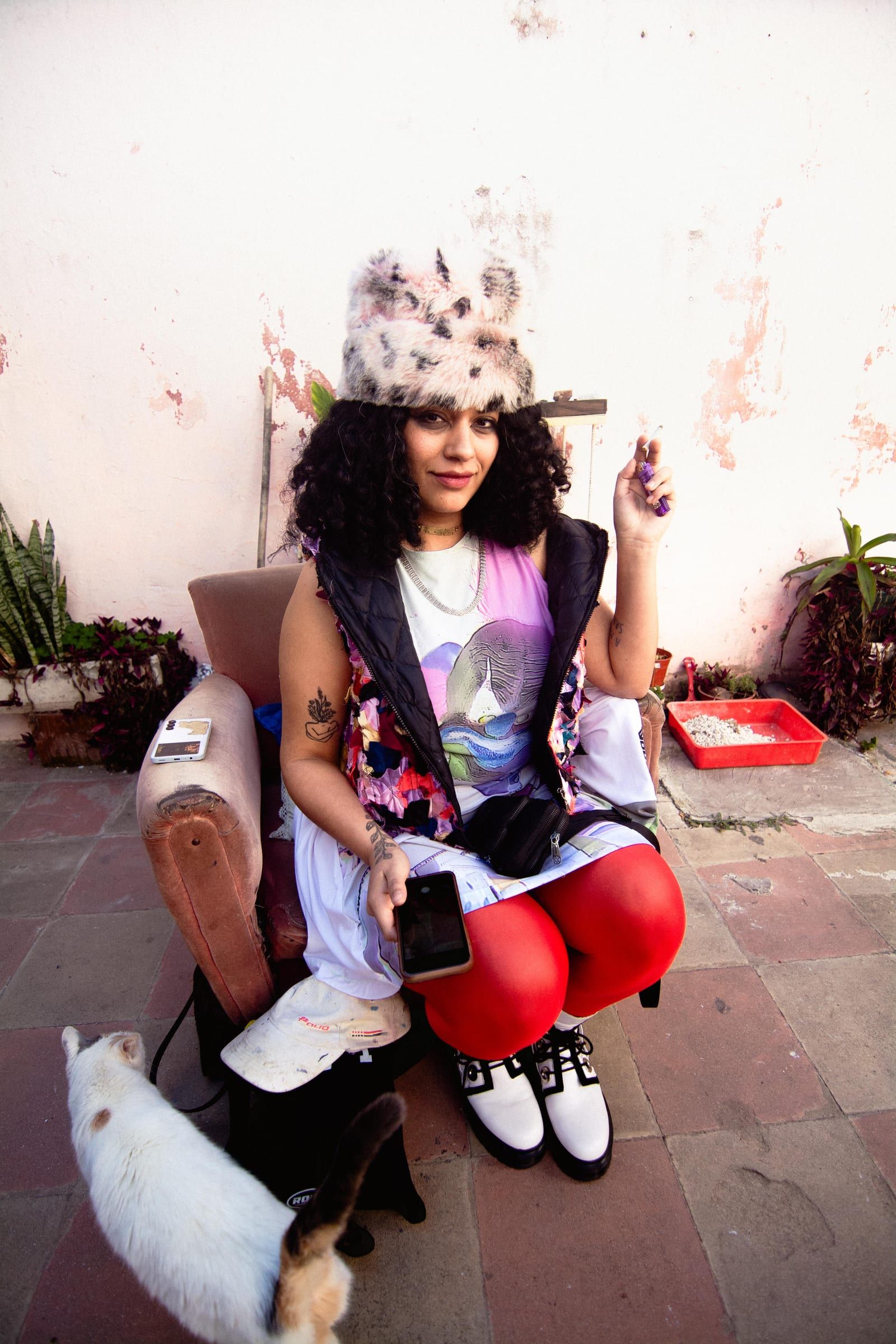 A person sits in the courtyard with a cat at their feet, in a fur hat and red boots.