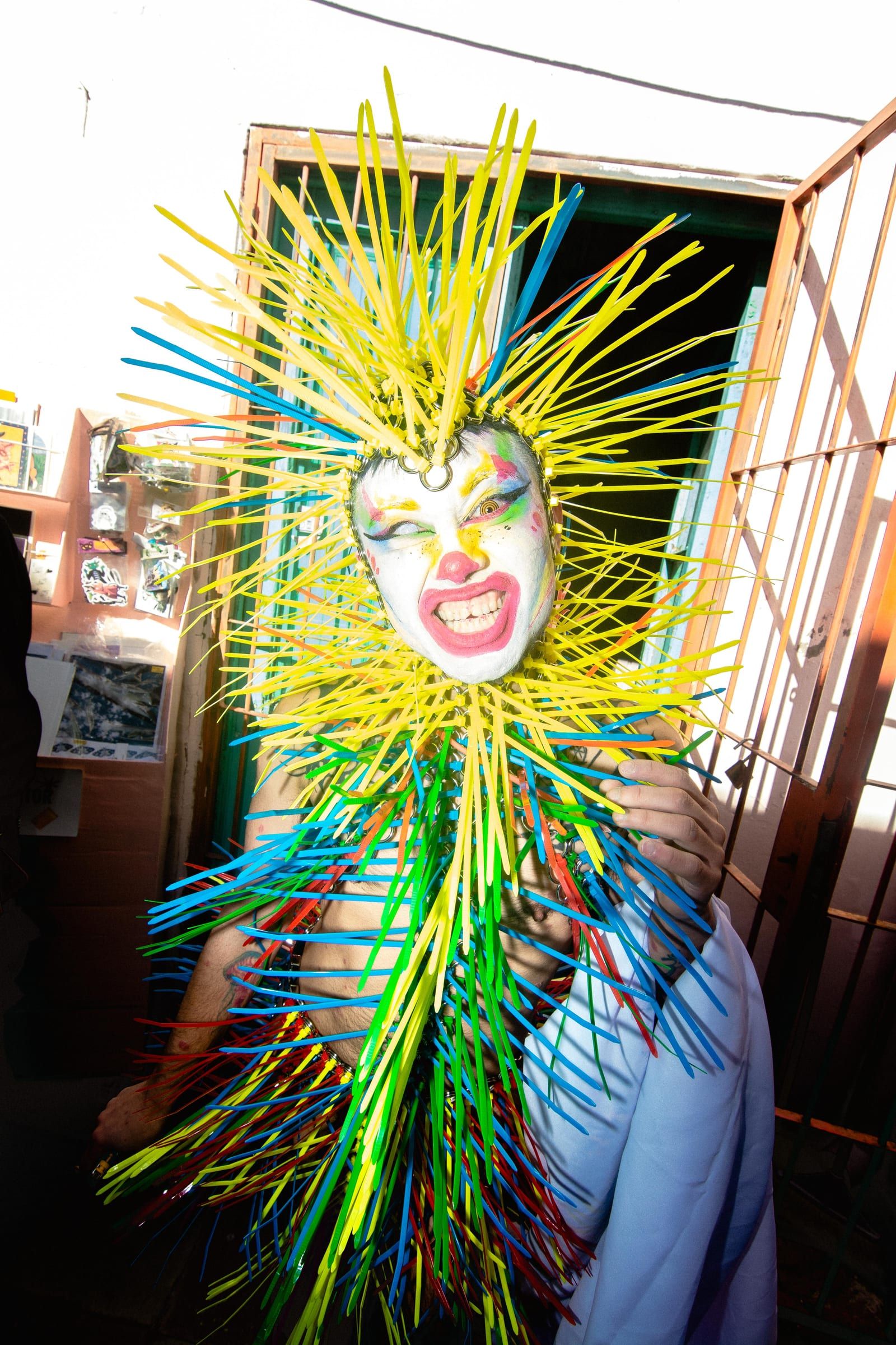 A figure in a yellow and green spiked costume waits in the courtyard, beside a railing.