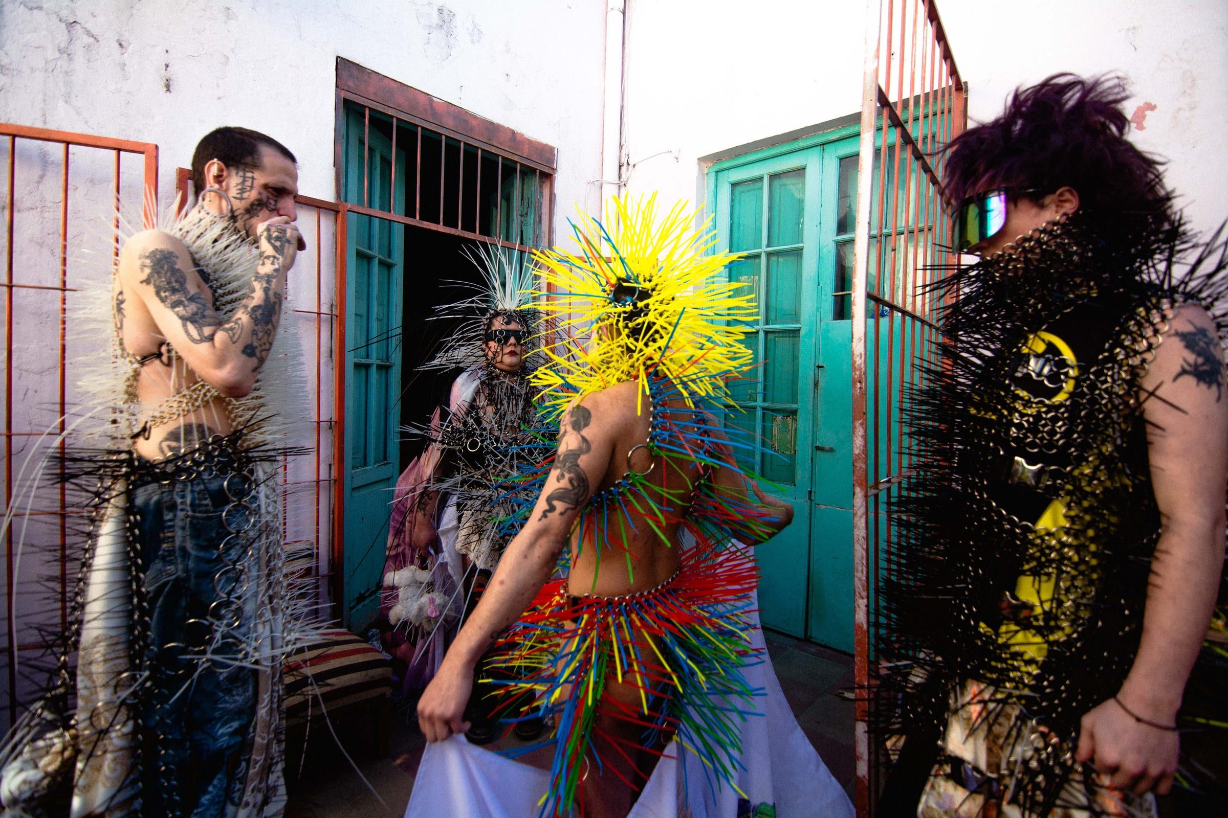 A group in coloured spiked costumes talks in the courtyard, in front of turquoise doors.