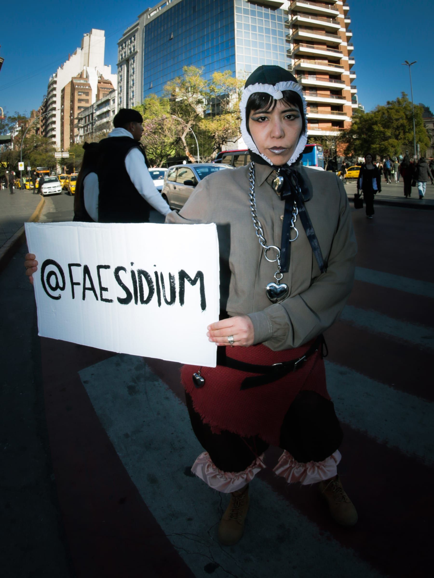 Una persona con gorro de piel sostiene un cartel escrito a mano que dice arroba Faesidium, parada en un cruce peatonal con edificios altos detrás.