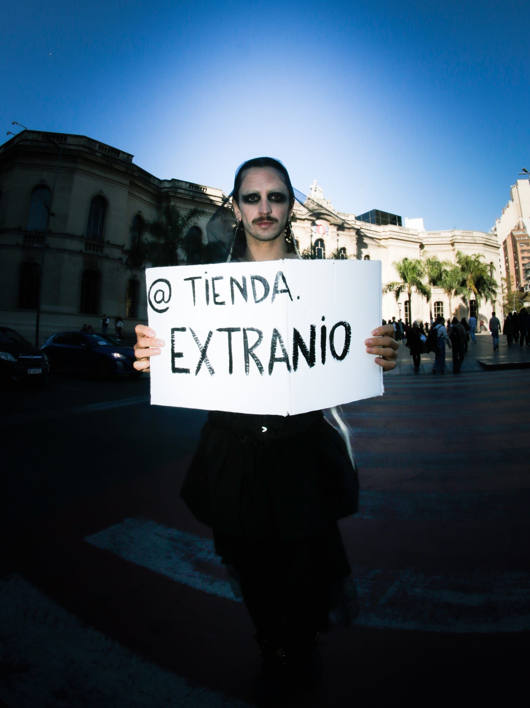 A person in dark glasses holds a handwritten sign reading at Tienda Extraño, with blue sky and a historic building behind.