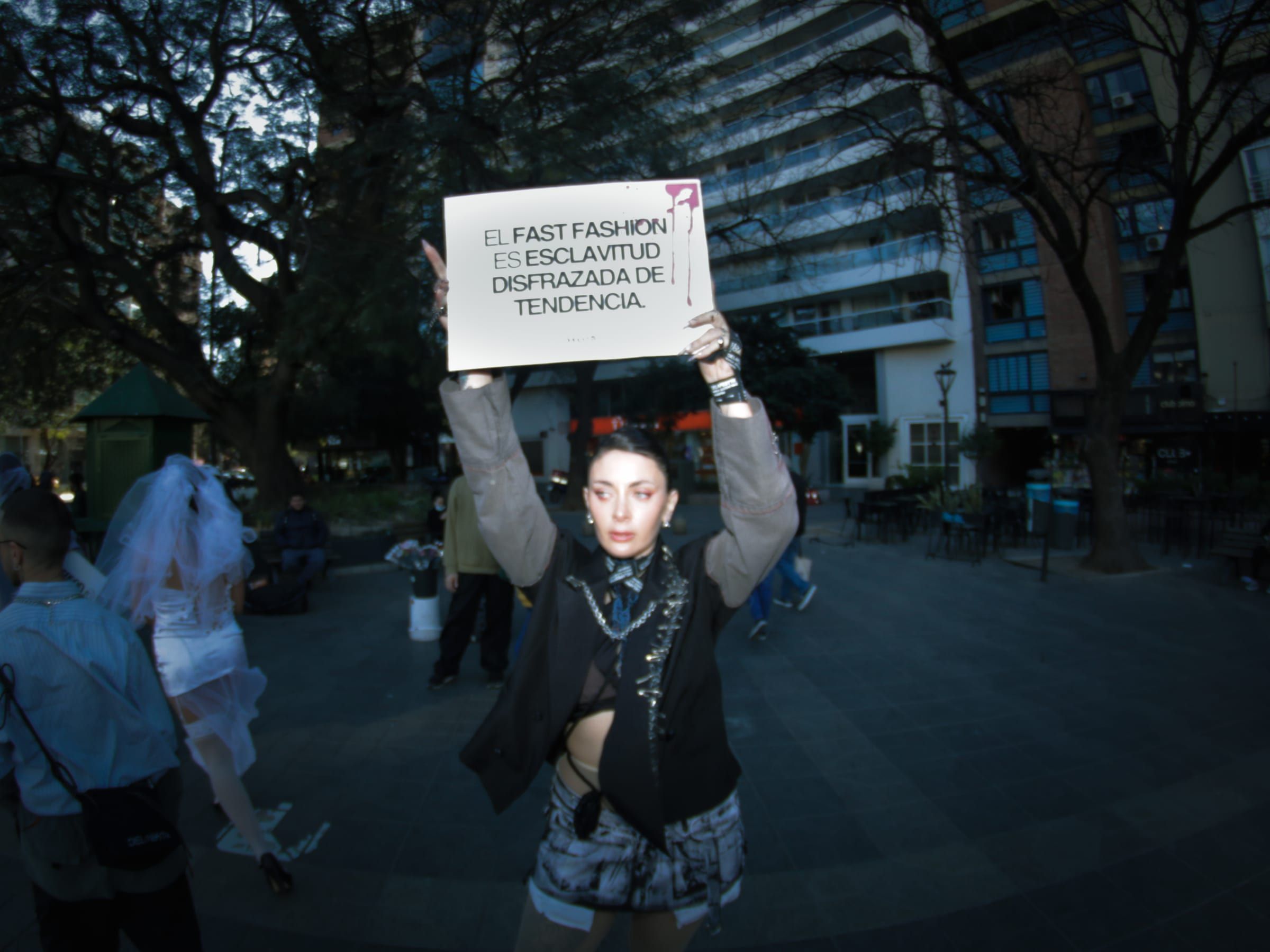 Una persona alza con las dos manos un cartel que dice: el fast fashion es esclavitud disfrazada de tendencia. Detrás, la plaza y sus árboles.