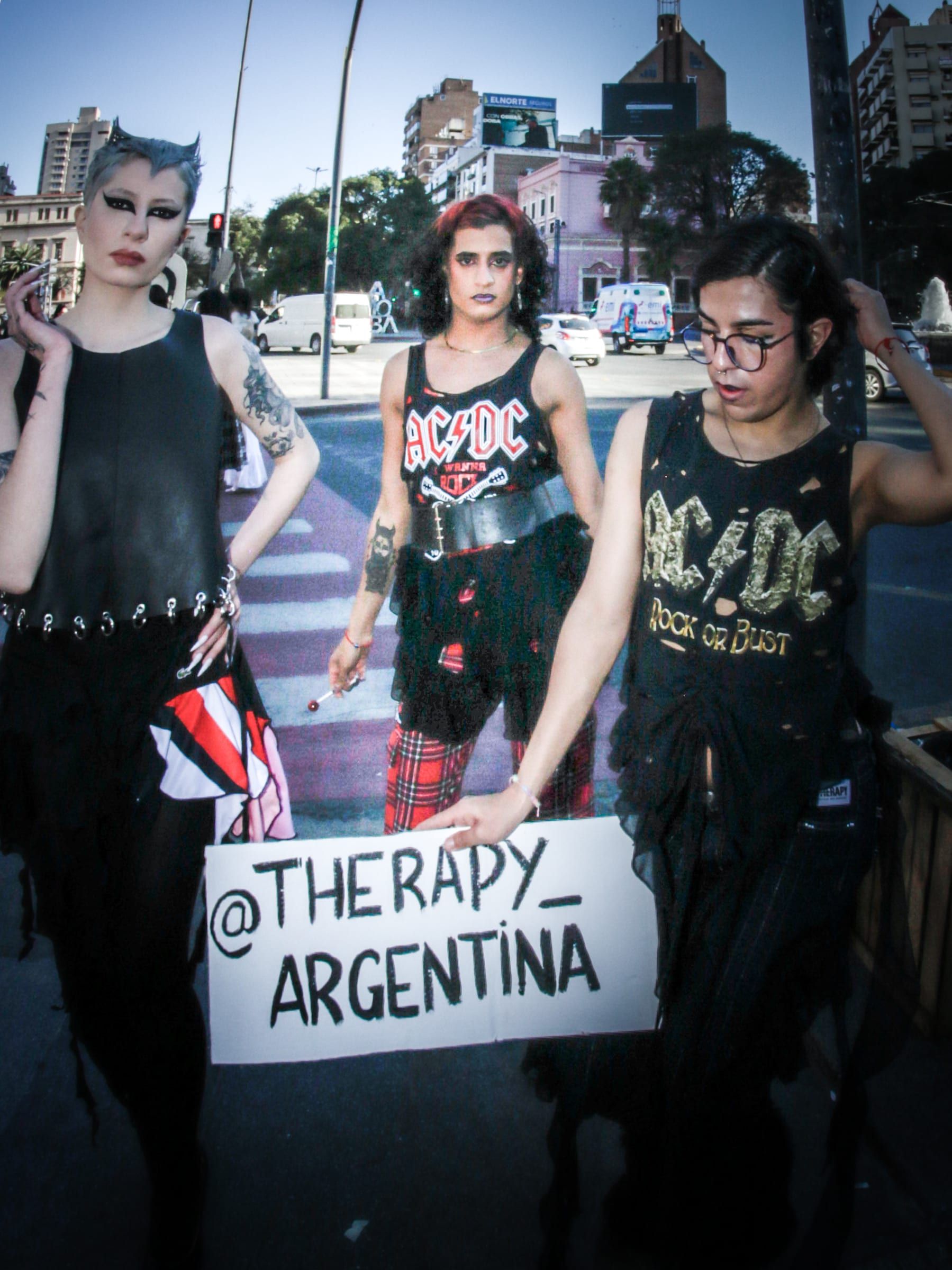 Tres personas vestidas de negro, con remeras de rock, sostienen un cartel que dice arroba Therapy Argentina, sobre una escalinata.