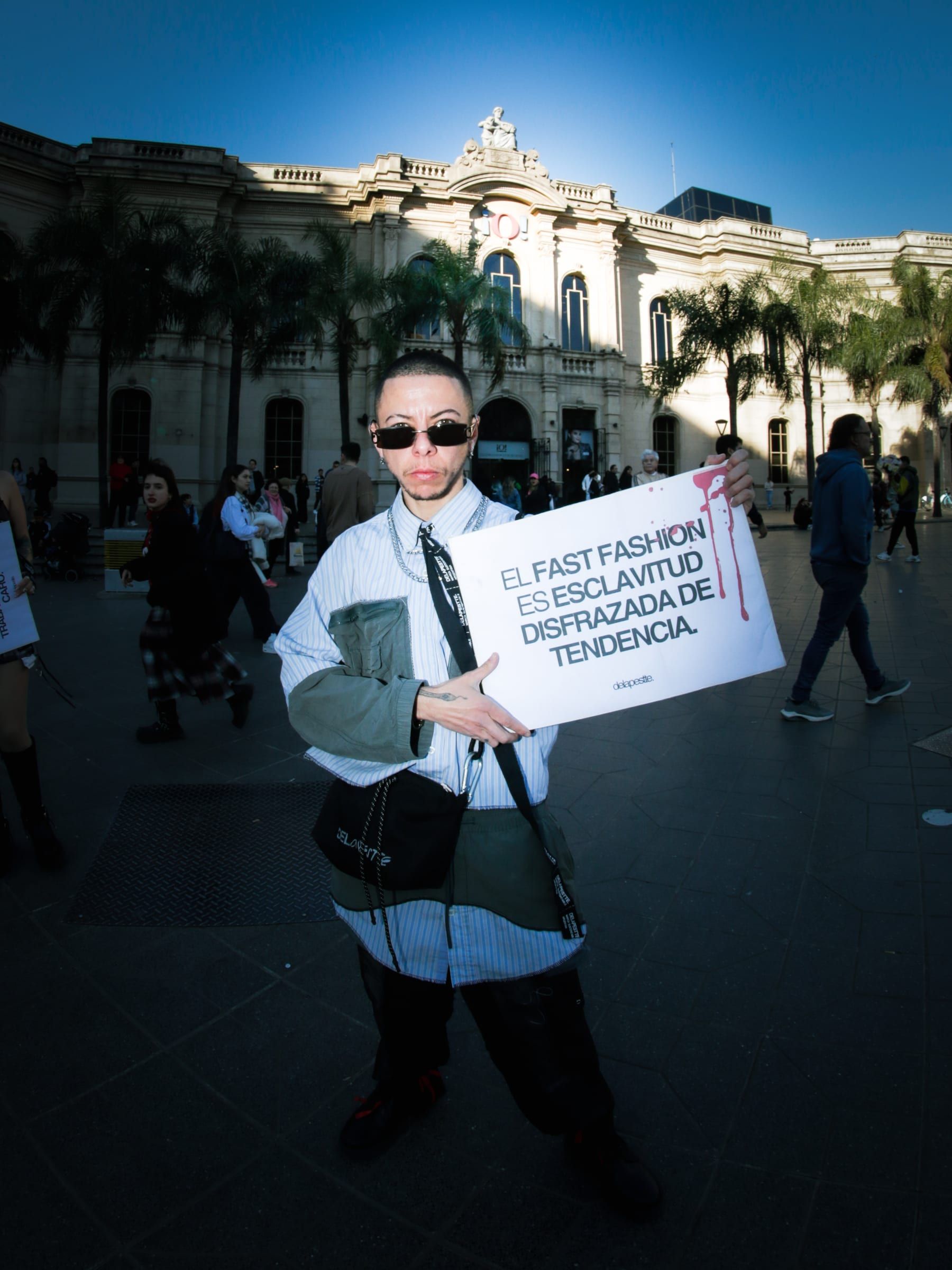 Una persona de camisa blanca y corbata sostiene el cartel sobre el fast fashion frente a un edificio histórico de la plaza.