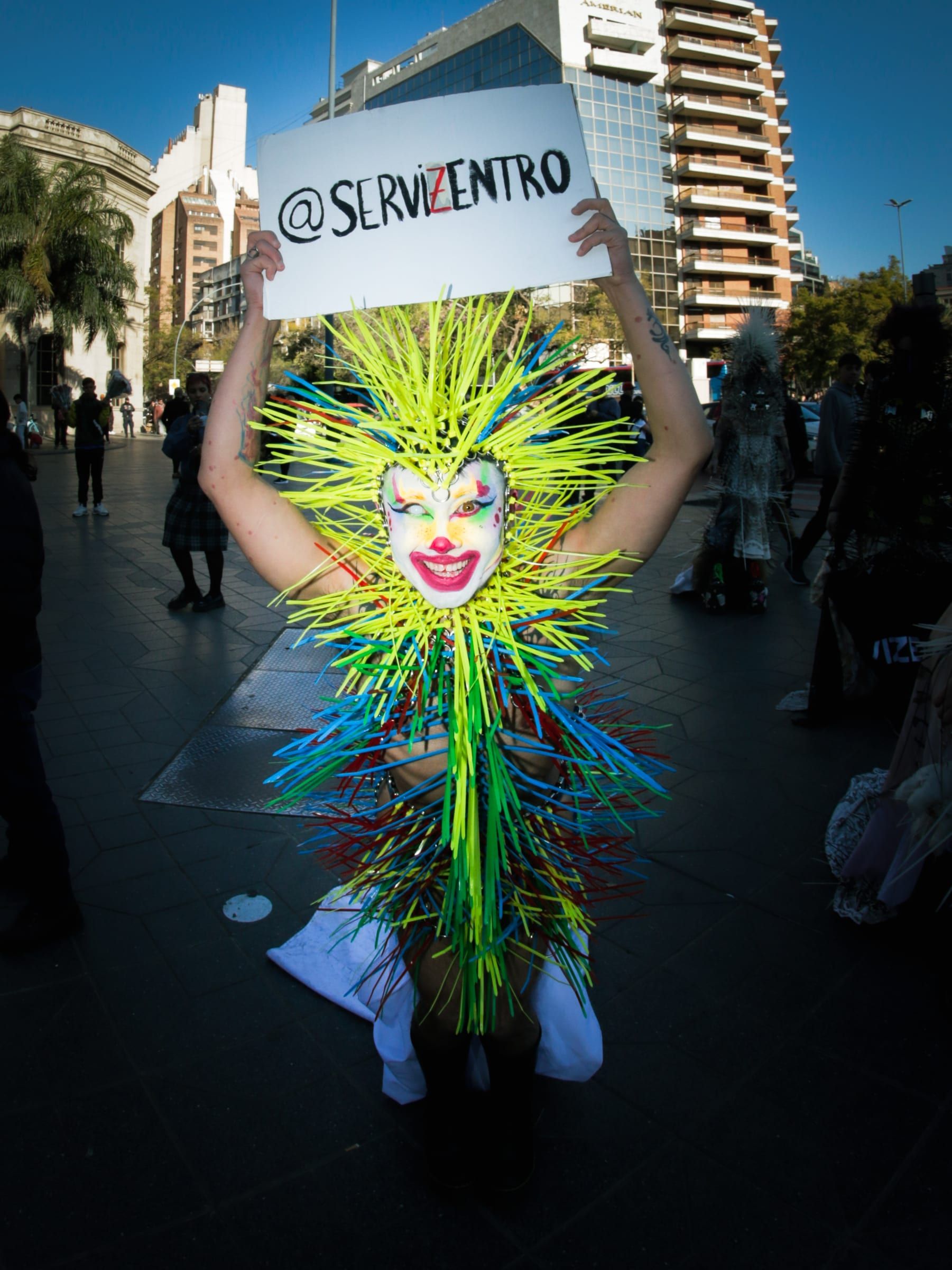 Una persona con un tocado de púas de colores alza sobre su cabeza un cartel que dice arroba Servizentro.