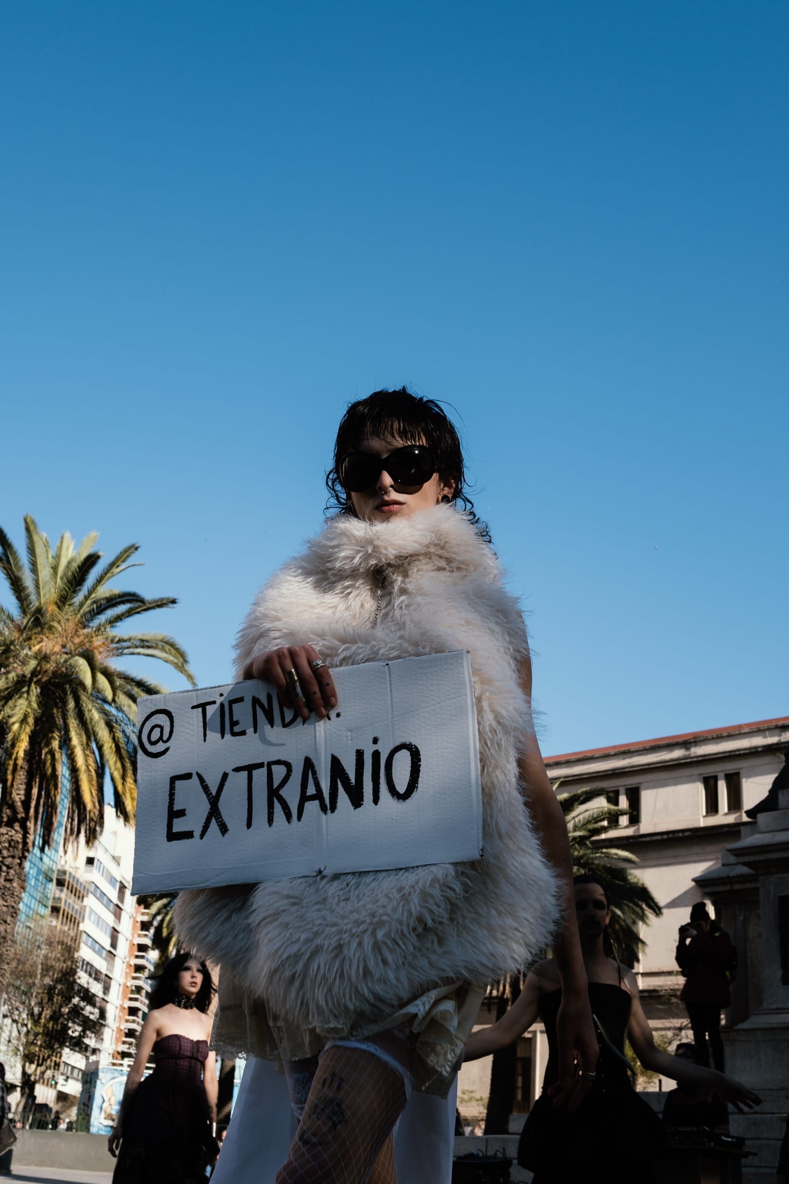 A person in a pale fur coat holds a sign reading at Tienda Extraño, among palm trees.