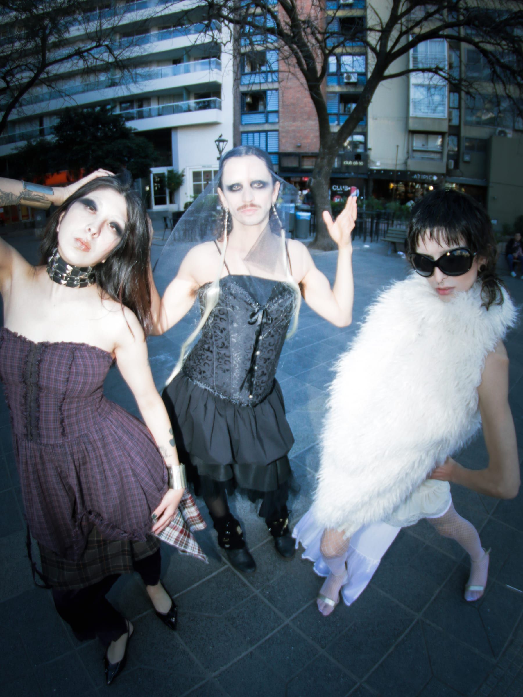 Three people pose in the square: two in corsets and skirts, one in a white feather coat.