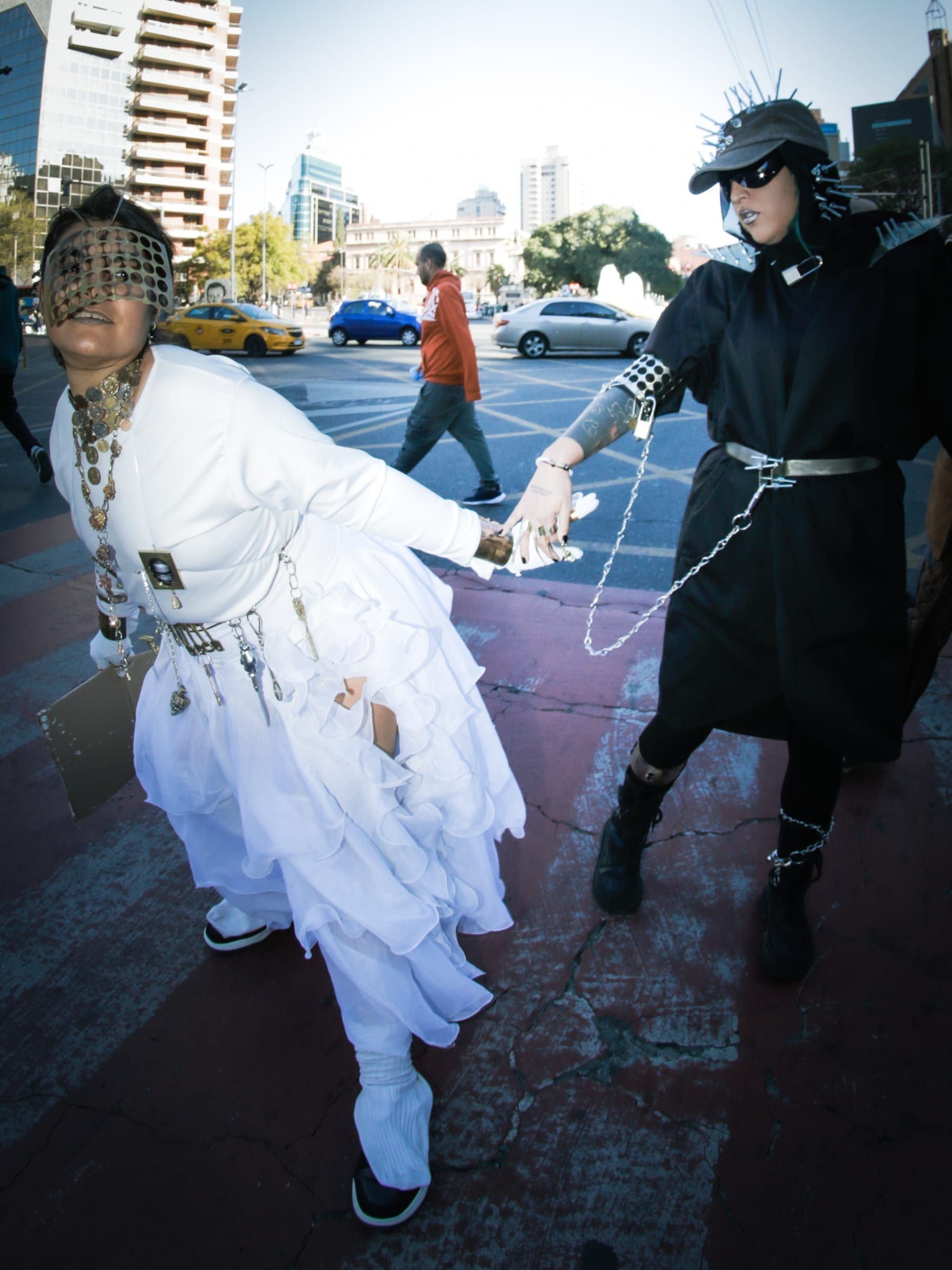 Two people cross the street hand in hand: one in white with a veil, the other in black with a mask.