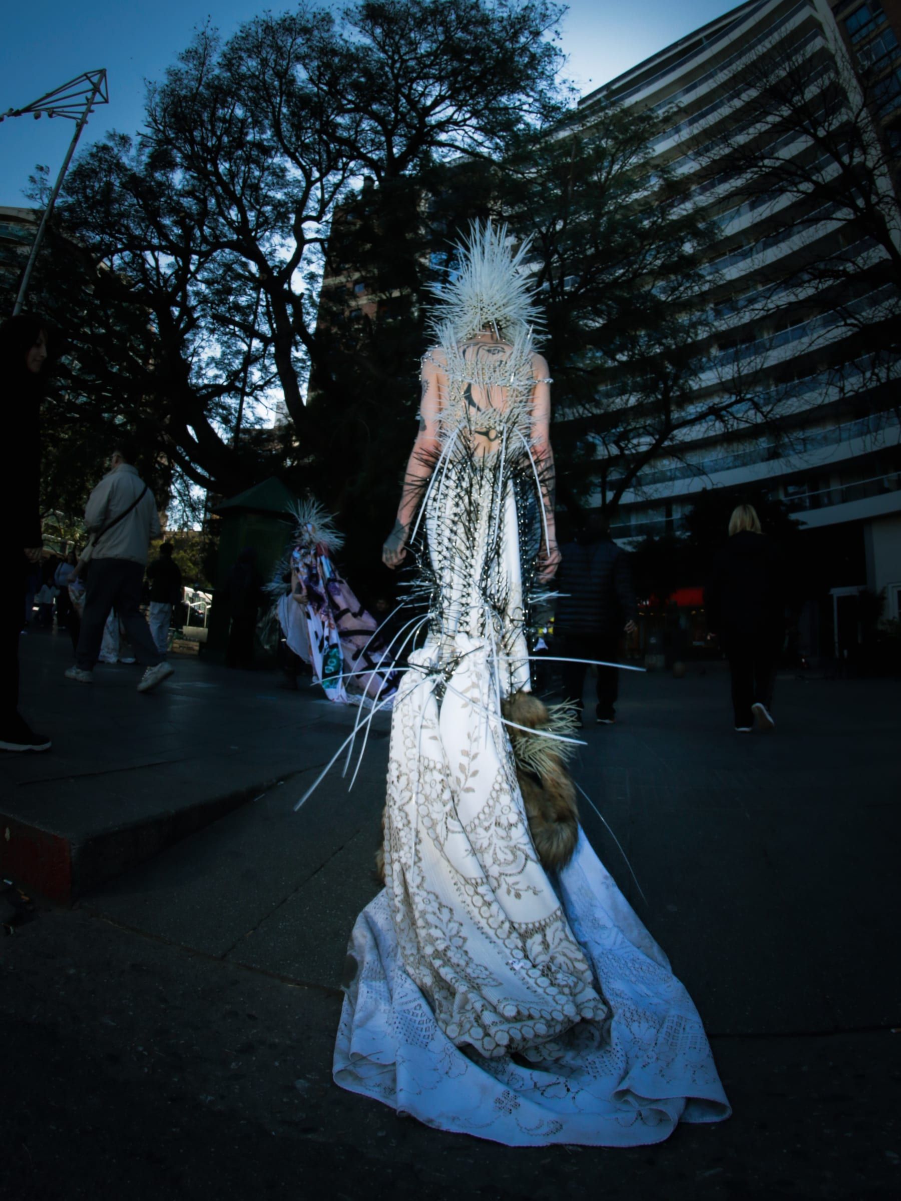 A person walks in a long white dress and a spiked headpiece, in front of an apartment building.