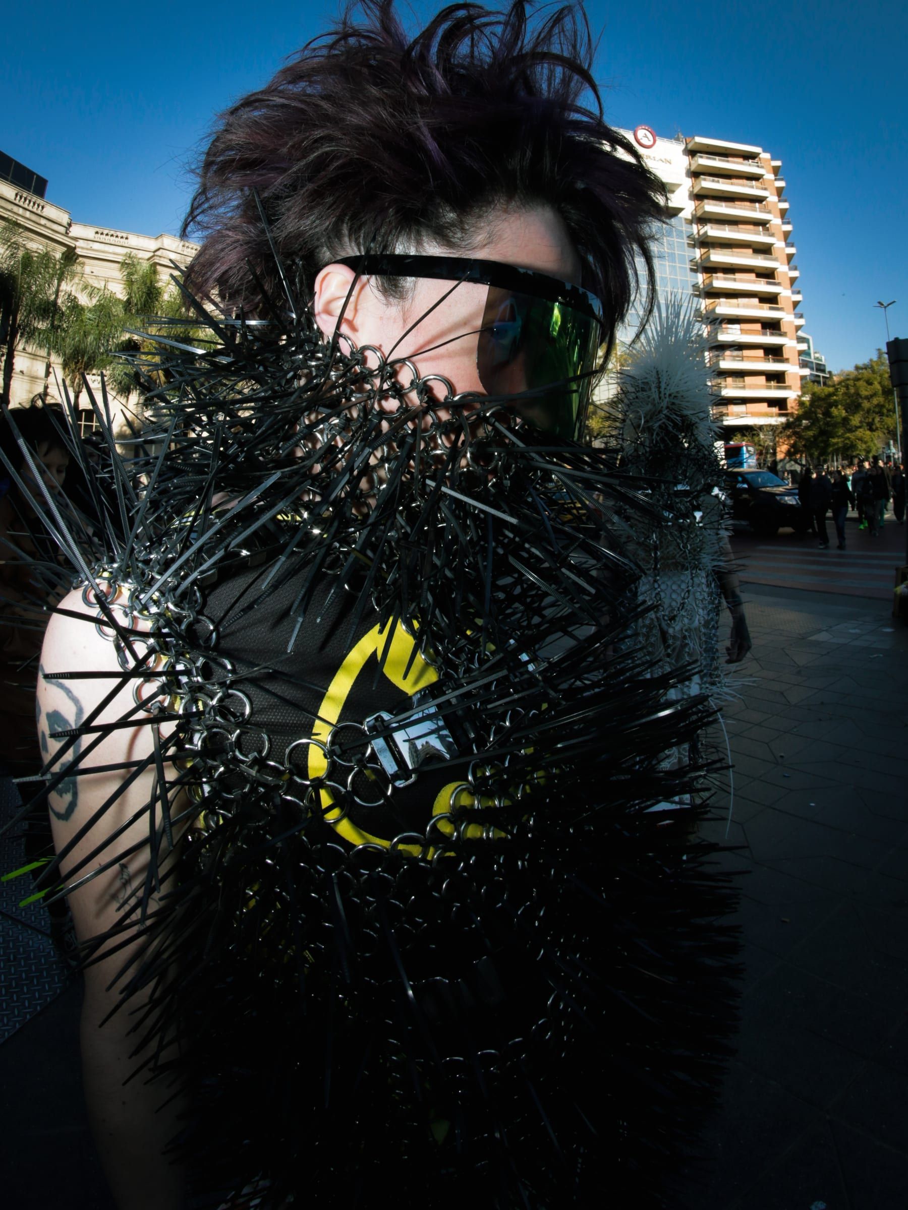 Close-up of a person with tousled hair, a dark visor and a costume covered in black spikes.