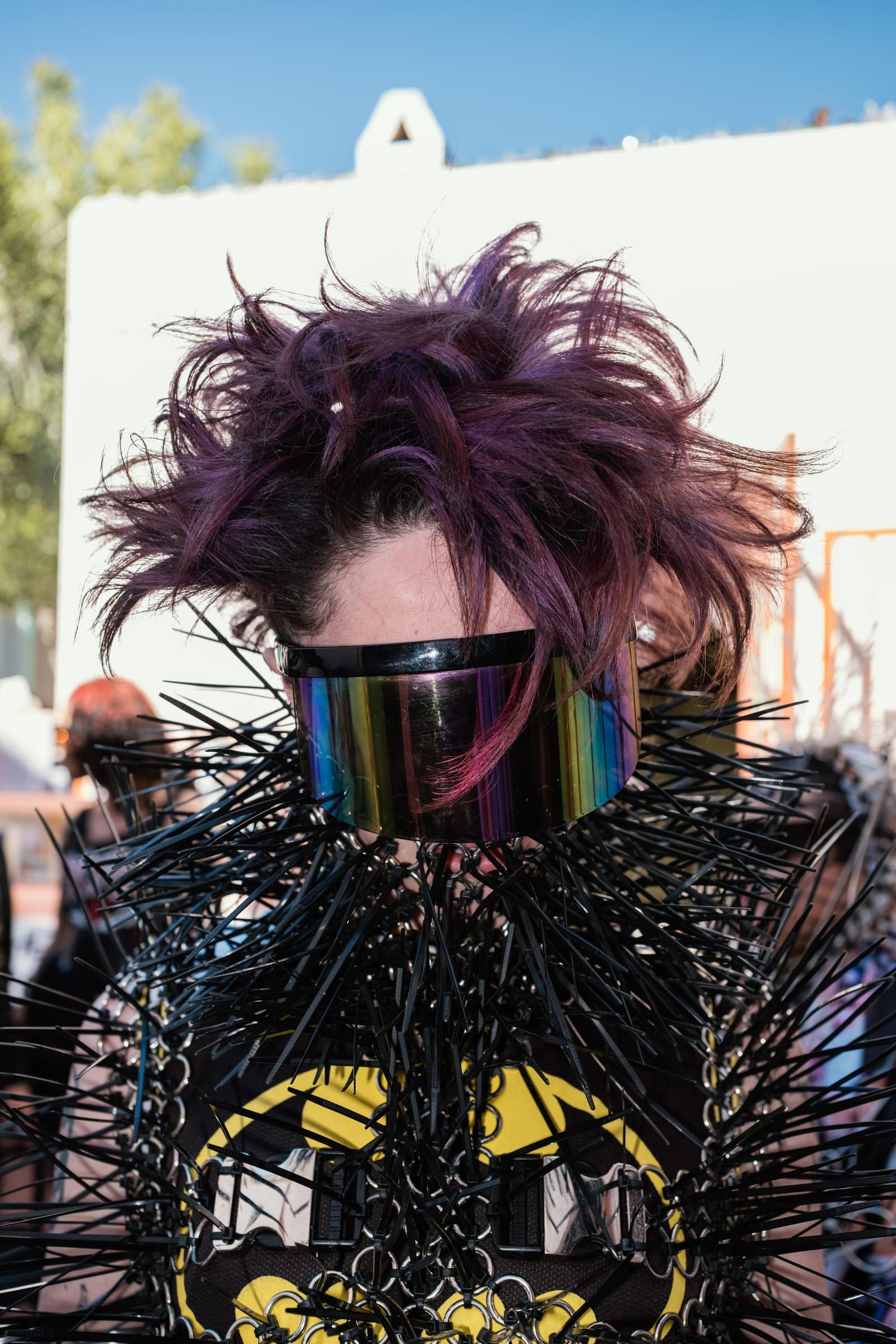 A person with purple hair and a mirrored visor wears a black spiked costume over a yellow shirt.