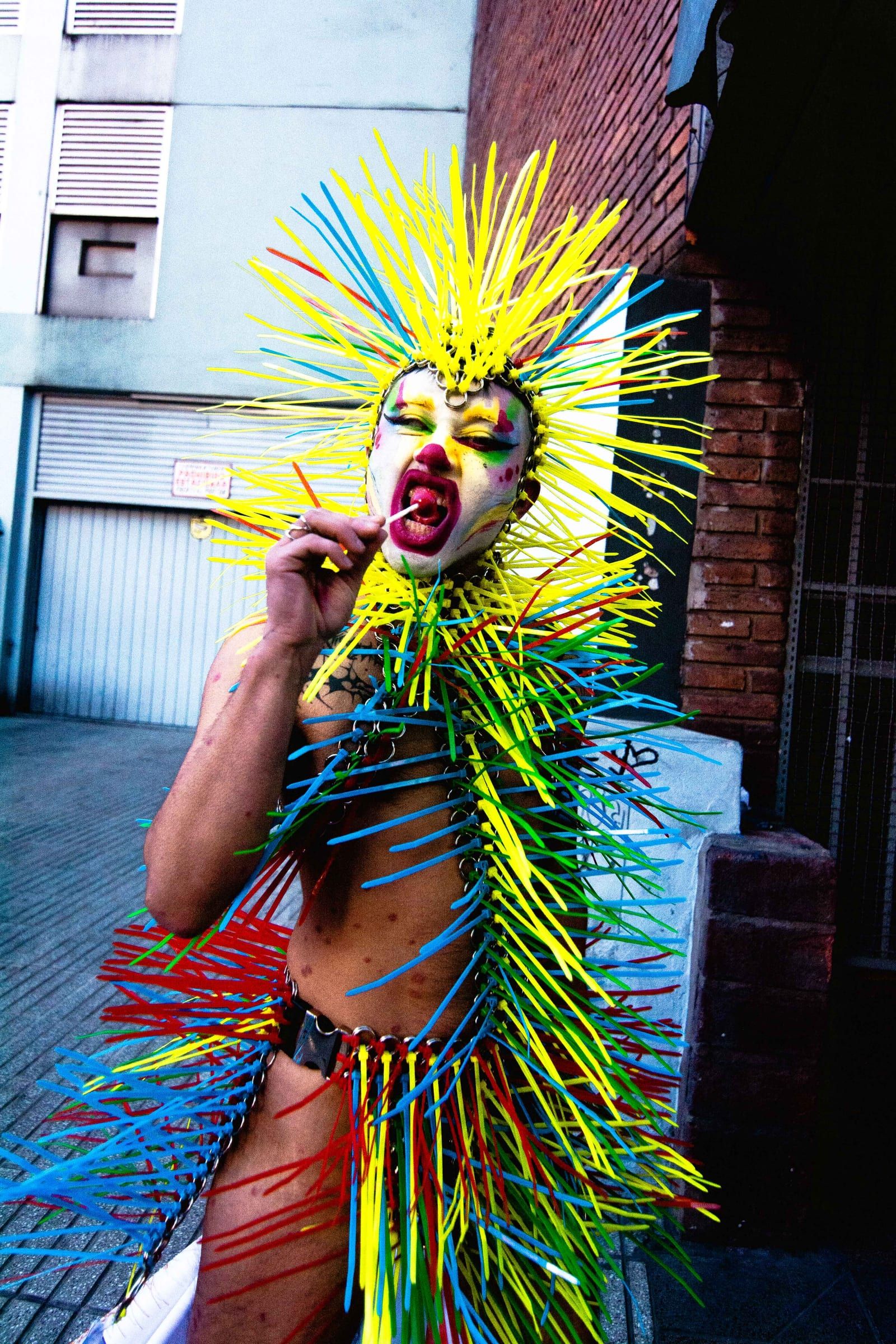 A person in a headpiece of coloured spikes shouts at the camera, in front of a grey shutter.