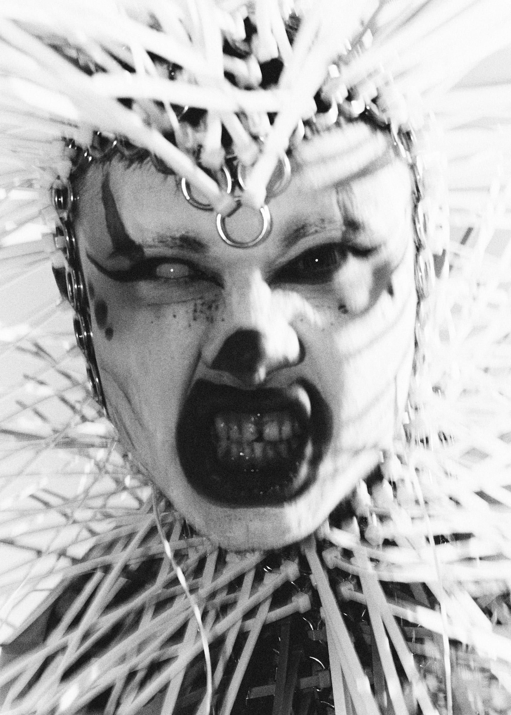 Black and white portrait: a made-up face with the mouth open, inside a headpiece of white spikes.